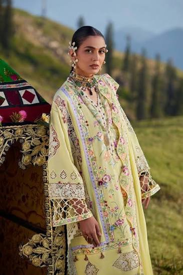 A woman stands outdoors in a grassy mountain landscape wearing a pale yellow embroidered traditional outfit with floral patterns and ornate lace detailing. A decorative camel or dromedary is partially visible beside her.