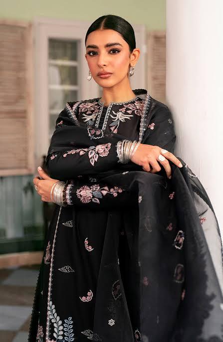A woman wearing a black embroidered traditional outfit with floral detailing and a matching sheer dupatta, posed against an outdoor architectural background.