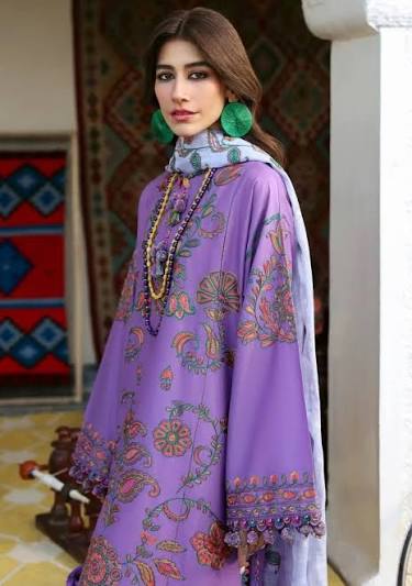 A woman wearing a lavender purple traditional outfit with colorful floral embroidery, a patterned scarf, and green earrings, standing in an indoor setting with a patterned rug and white pillar visible.