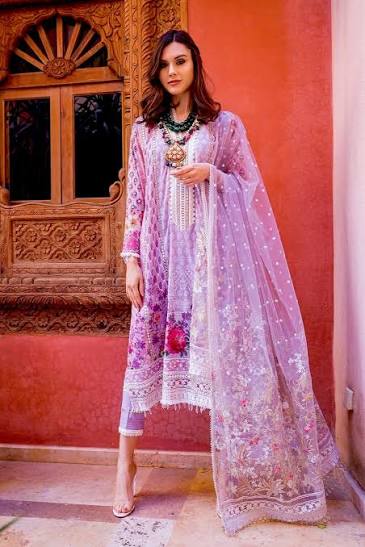 A woman models a traditional three-piece salwar kameez suit in light purple with white floral embroidery and a matching sheer dupatta, set against a warm terracotta wall and carved wooden door.