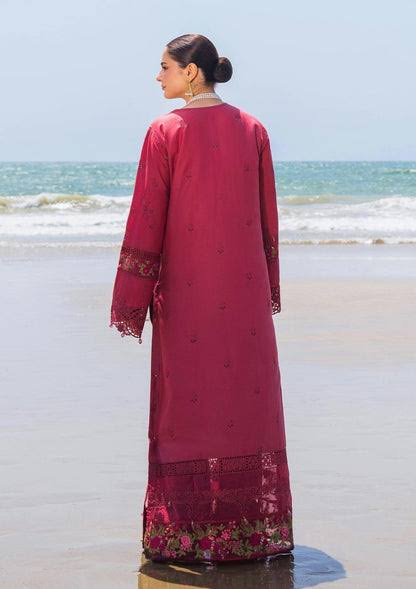 A woman stands on a beach facing the ocean, wearing a long magenta-pink traditional dress with lace cuffs and a floral lace hem.