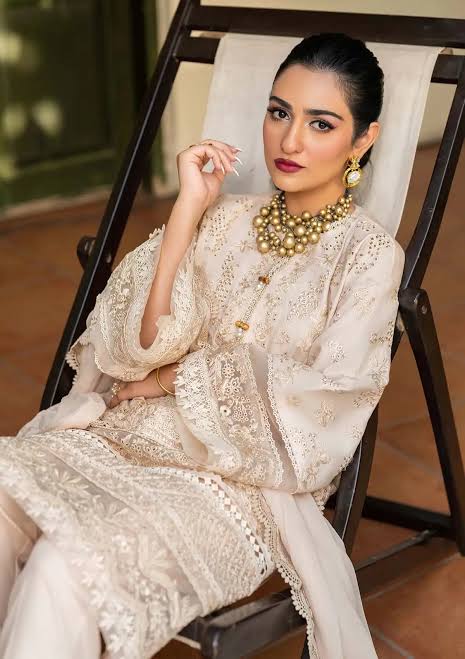 A woman wearing a cream-colored embroidered traditional outfit sits in a dark wooden lounge chair. The outfit features intricate floral embroidery and sheer sleeves, paired with a matching sheer dupatta.