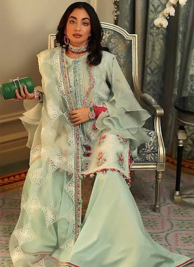 A woman seated in an ornate chair wearing a pale mint green traditional outfit with white embroidered lace details and pink floral accents. She holds a green clutch in a decorated indoor setting with patterned carpet and drapery.