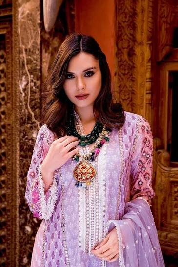 A woman models a traditional three-piece light purple and white embroidered salwar kameez set with a matching dupatta, styled with a statement green and gold necklace, set against an ornate golden carved background.