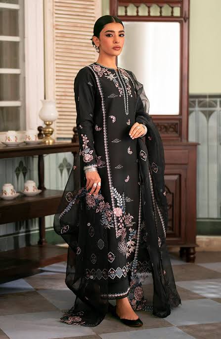A woman models a black three-piece traditional outfit with pink and white floral embroidery, featuring a long kurta, matching trousers, and a sheer dupatta, set against an ornate indoor background.