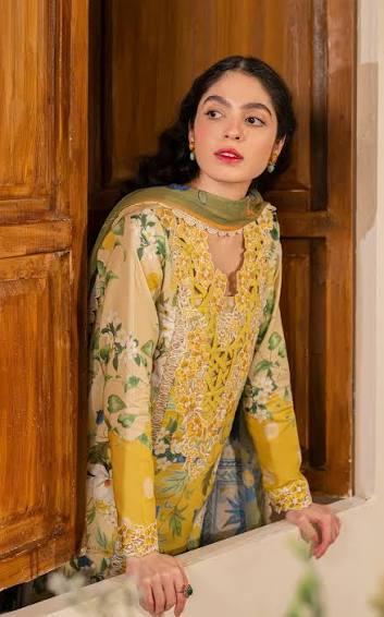 A woman wearing a yellow and beige printed traditional outfit with green accents, standing in front of a rustic wooden door.