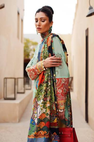 A woman wearing a colorful, highly patterned traditional long dress with a matching dupatta, standing in a sunlit outdoor alleyway. She is holding a bright red handbag.