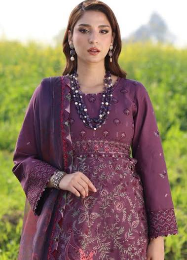 A woman wearing a purple embroidered traditional outfit with a matching dupatta, standing outdoors against a blurred green field background.