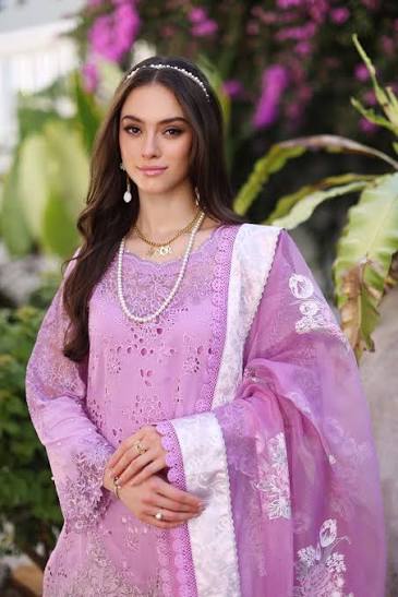 A woman wearing a lavender purple embroidered traditional outfit with a white border and matching sheer dupatta, styled with pearl necklaces and a silver headpiece, set against a soft-focus background of green foliage and purple flowers.