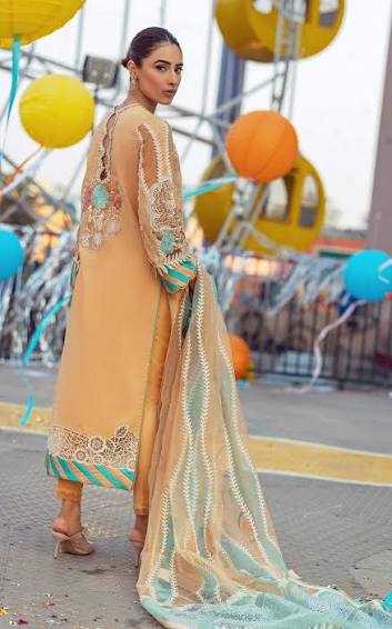 A woman modeling a peach-colored traditional outfit with intricate embroidery and a long sheer dupatta, photographed outdoors with colorful balloons and a blurred event background.