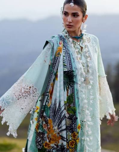 A woman wearing a traditional embroidered outfit with a mint green and white lace outer layer, a patterned teal dupatta, and a matching printed inner garment, photographed outdoors against a blurred mountainous background.