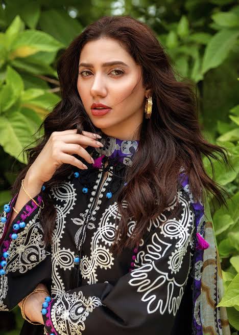 A woman with long, wavy dark brown hair is shown in a lifestyle setting, wearing a black embroidered top with white, purple, and blue patterns, set against a background of lush green foliage.