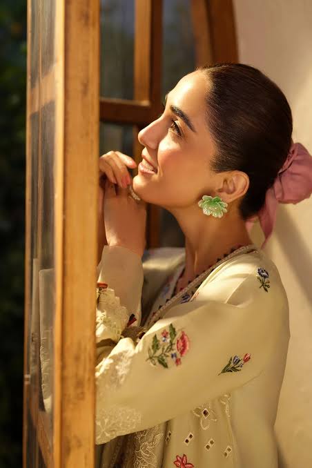 A woman in profile wearing a cream-colored embroidered traditional outfit and a pink hair bow, standing next to a wooden window frame.