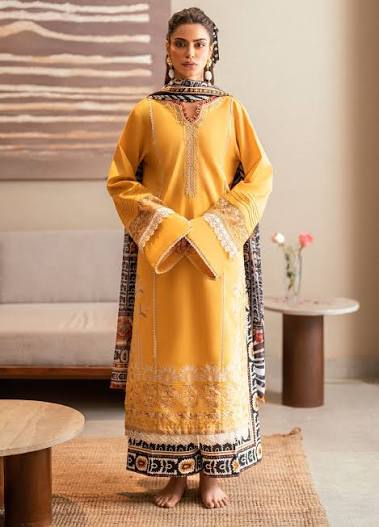 A woman stands barefoot indoors wearing a long yellow traditional outfit with ornate embroidery, patterned cuffs, and a matching patterned shawl draped over her shoulders. The room has neutral walls, a sofa, and small tables.
