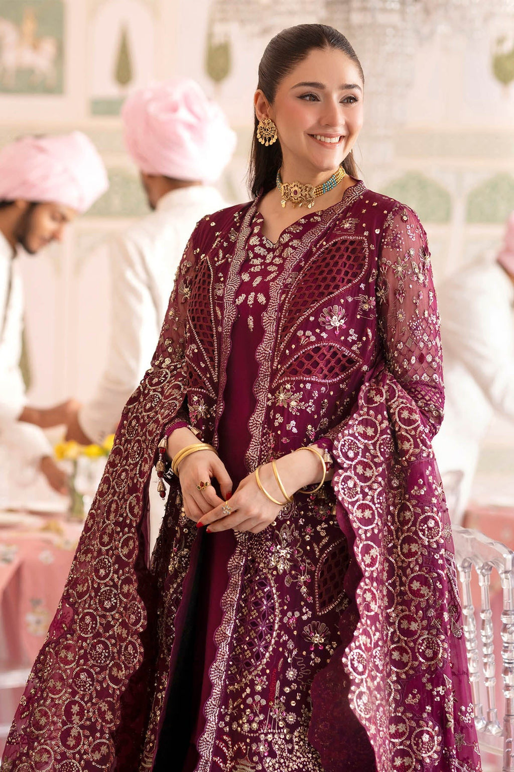 A woman wearing an ornate deep magenta embroidered traditional outfit with gold detailing, standing in a decorated indoor setting with blurred figures in the background.