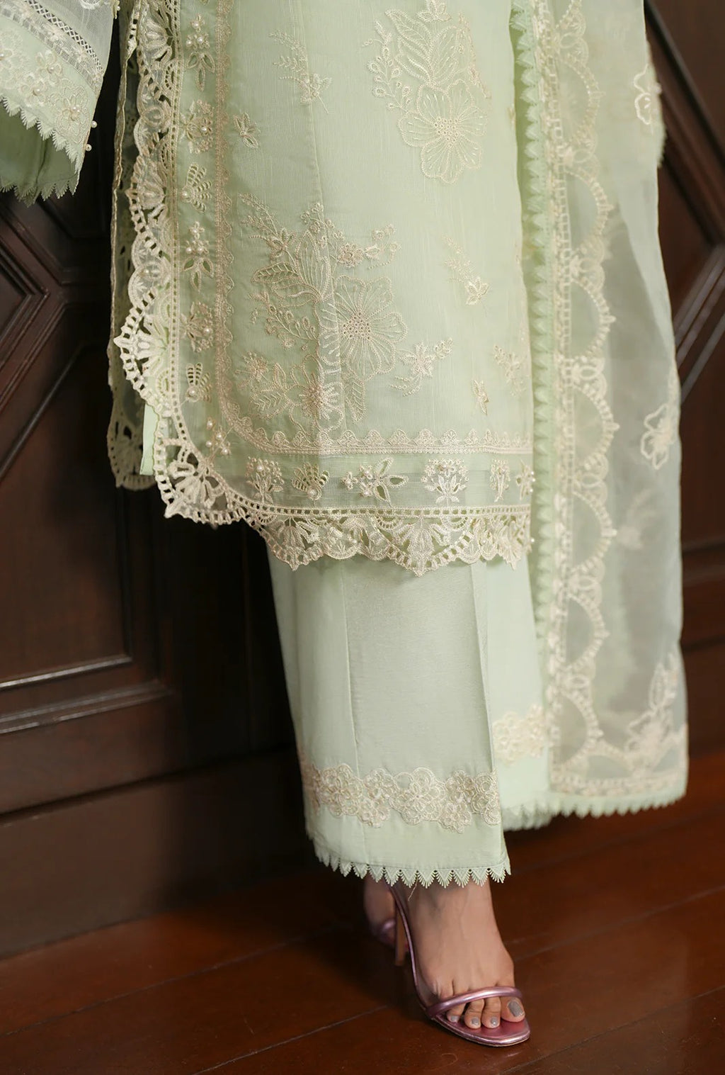 A close-up of a person wearing a light mint green traditional outfit with intricate cream embroidery and lace borders, paired with metallic pink high-heeled sandals, standing on a dark wood floor.