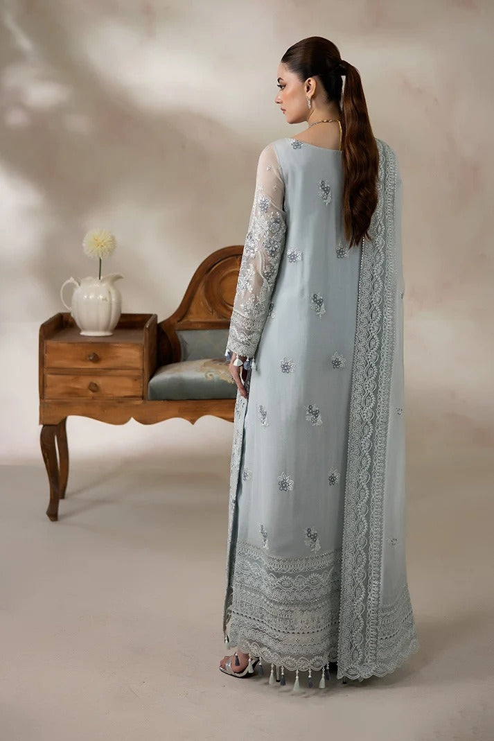 A woman stands with her back to the camera wearing a light blue-gray traditional outfit with intricate silver embroidery and a matching sheer dupatta. She is posed in front of a rustic wooden bench and a small side table with a vase, set against a warm beige studio backdrop.