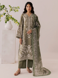 A woman models a three-piece olive green ethnic suit with intricate white floral embroidery, matching trousers, and a long embroidered dupatta, set against a neutral studio background with a decorative plant on a pedestal.