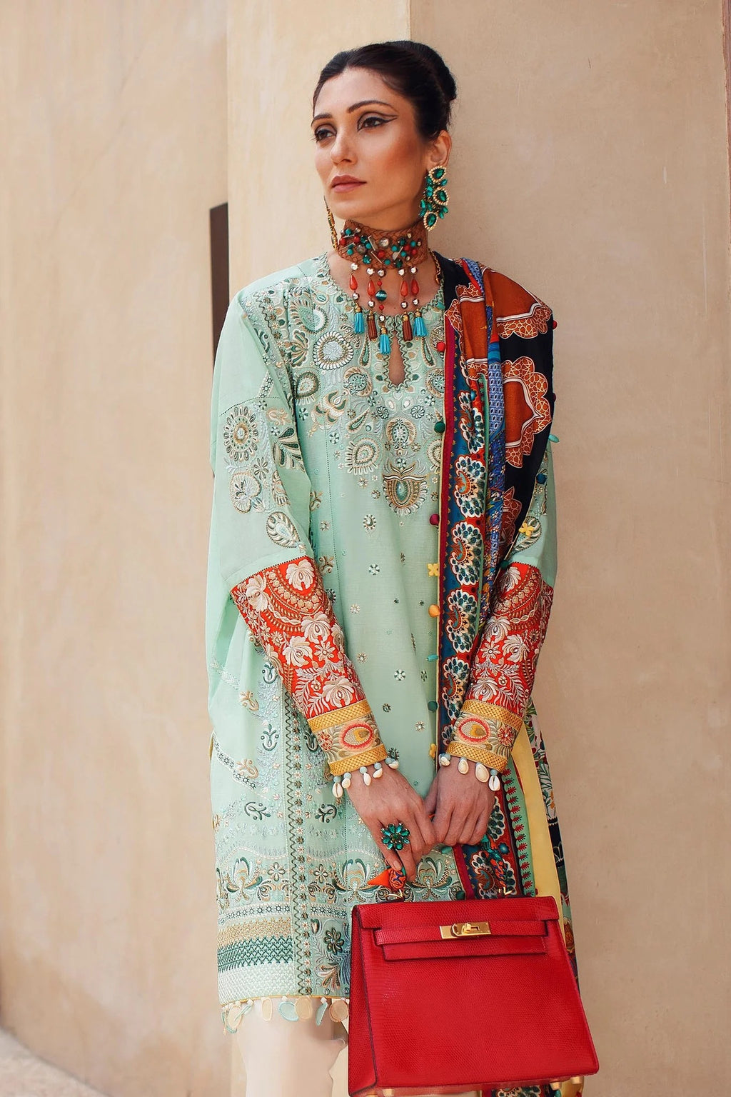 Woman in traditional embroidered outfit with a red handbag against a beige wall.