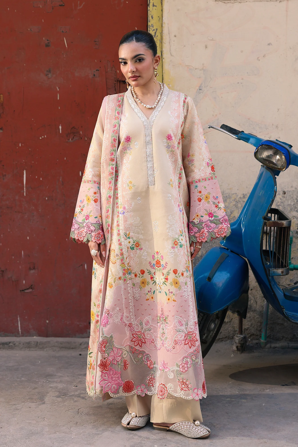 A woman models a traditional long beige and pink floral embroidered kurta with a matching sheer jacket, standing next to a blue scooter in front of a rustic red and beige wall.