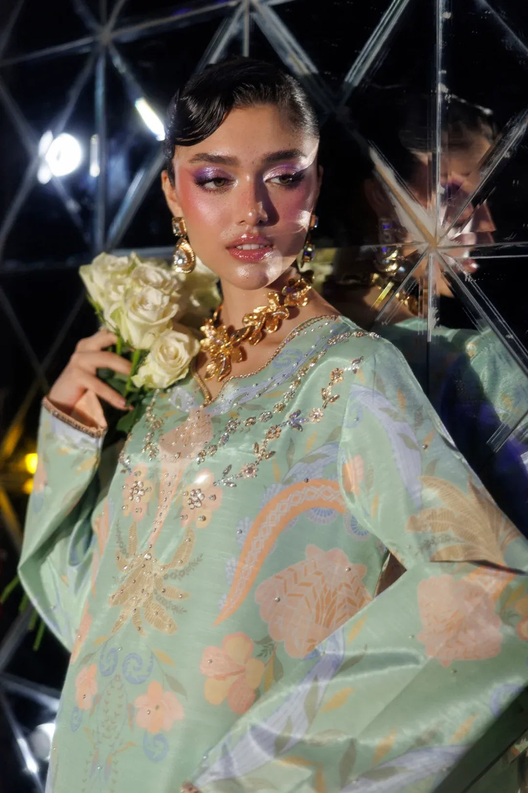 A model wearing a light sage green embroidered kurta with floral and paisley patterns in gold, pink, and purple, accessorized with a chunky gold chain necklace and holding a bouquet of white roses in front of a mirrored geometric wall.