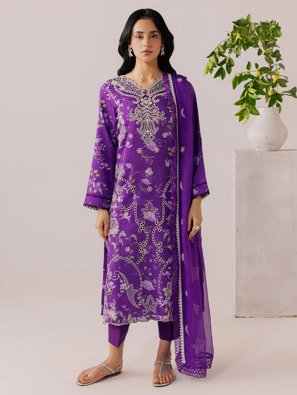 A woman modeling a purple traditional kurta set with intricate silver embroidery, matching pants, and a sheer dupatta, standing in a studio with a potted plant in the background.