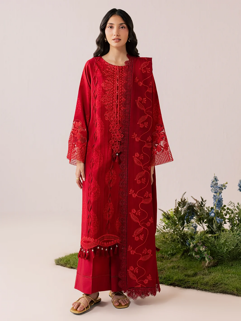A woman modeling a long red embroidered kurta with matching pants in a studio setting with a light beige background and decorative plants at the bottom right.