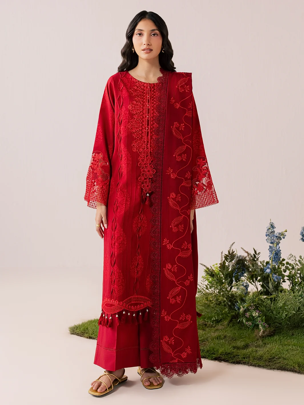 A woman modeling a long red embroidered kurta with matching pants in a studio setting with a light beige background and decorative plants at the bottom right.