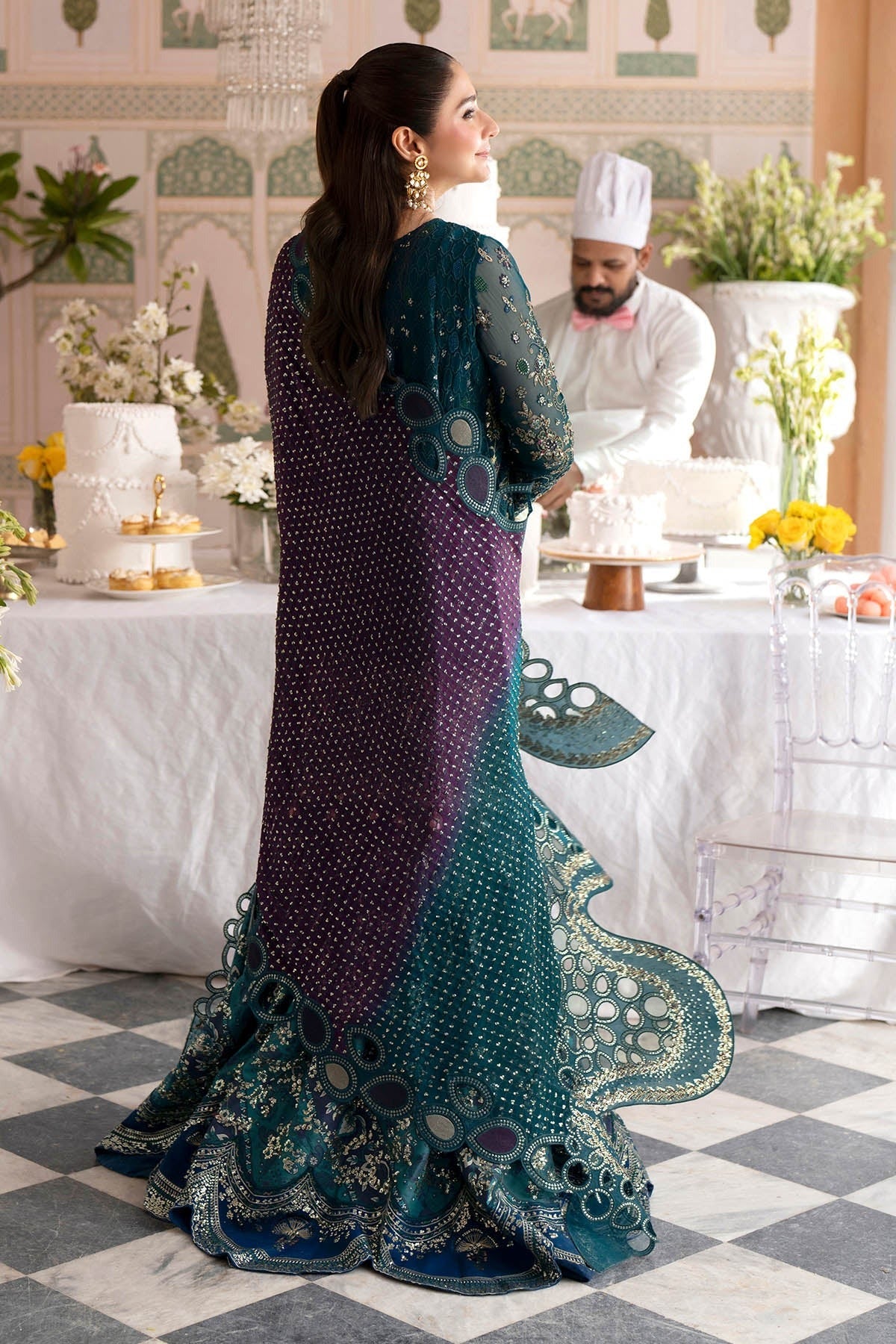 A woman wearing a dark teal and purple embroidered traditional gown with a matching sheer dupatta, photographed from behind in an elegant event setting with tables, cakes, and a chef in the background.