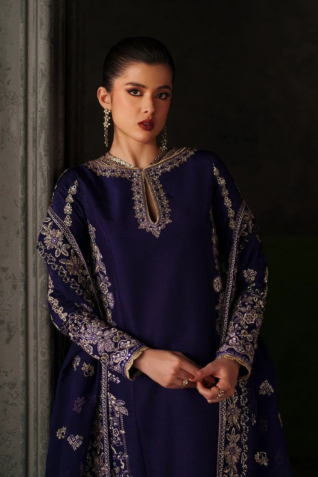 A woman wearing a deep navy blue traditional outfit with ornate gold embroidery, standing against a dark background.