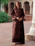 A woman stands outdoors in a courtyard wearing a long, dark brown embroidered traditional outfit with ornate floral detailing and a matching dupatta. The background shows a stone building with arches and a brick-paved walkway.
