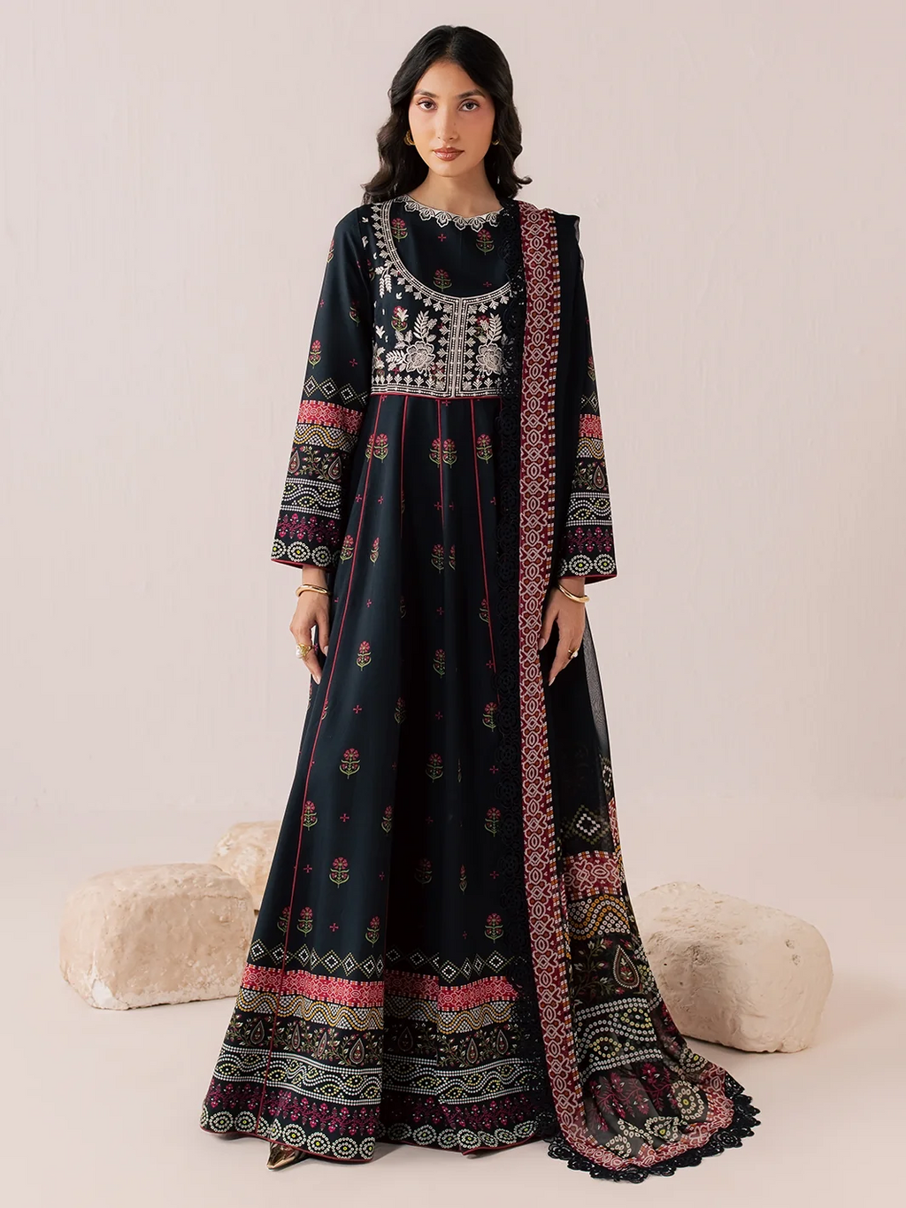 A woman models a long black embroidered Anarkali gown with a matching dupatta, featuring intricate silver and pink floral patterns, set against a neutral beige background with decorative pillows.