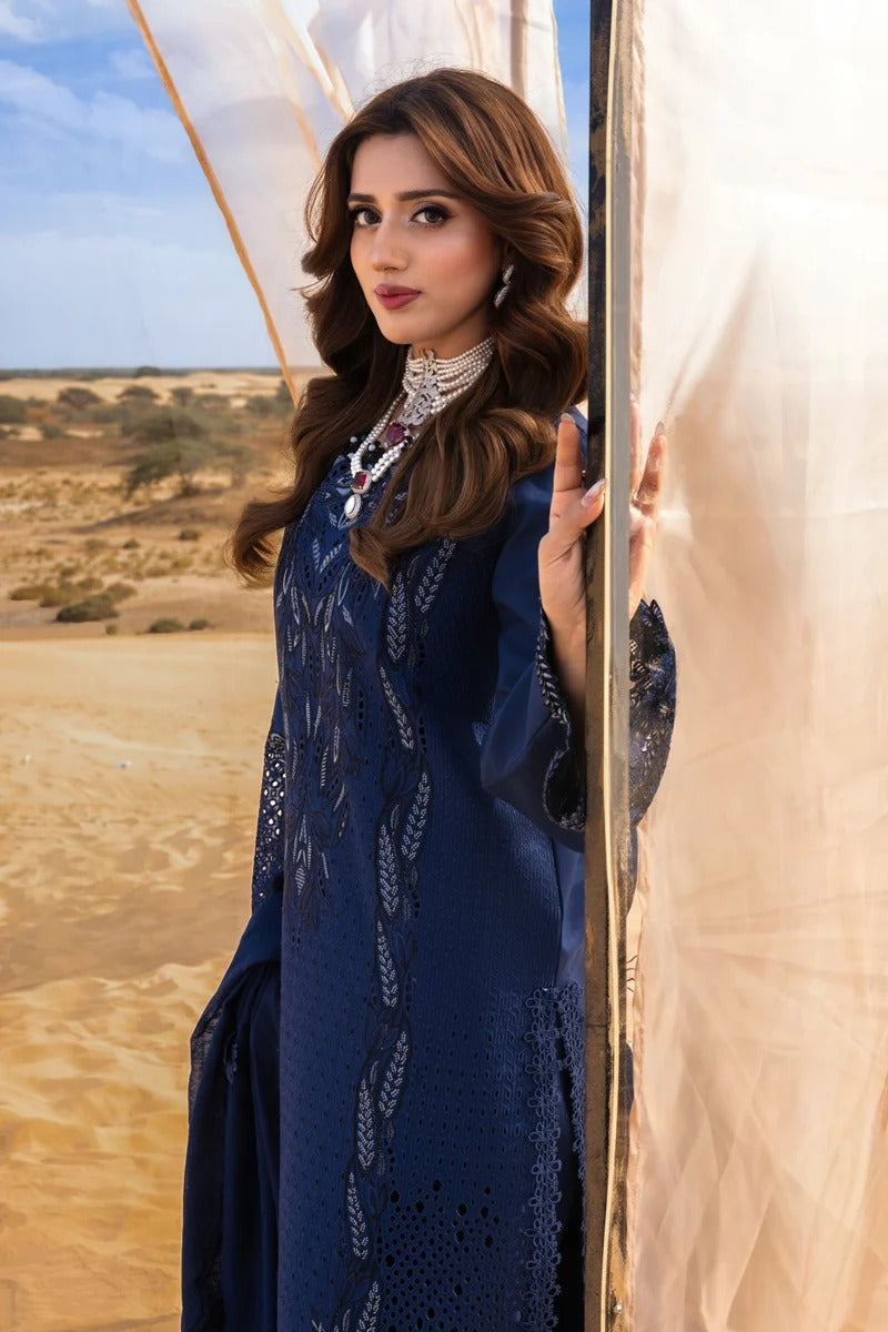 A woman wearing a deep navy blue embroidered traditional outfit stands beside a white tent in a desert landscape.