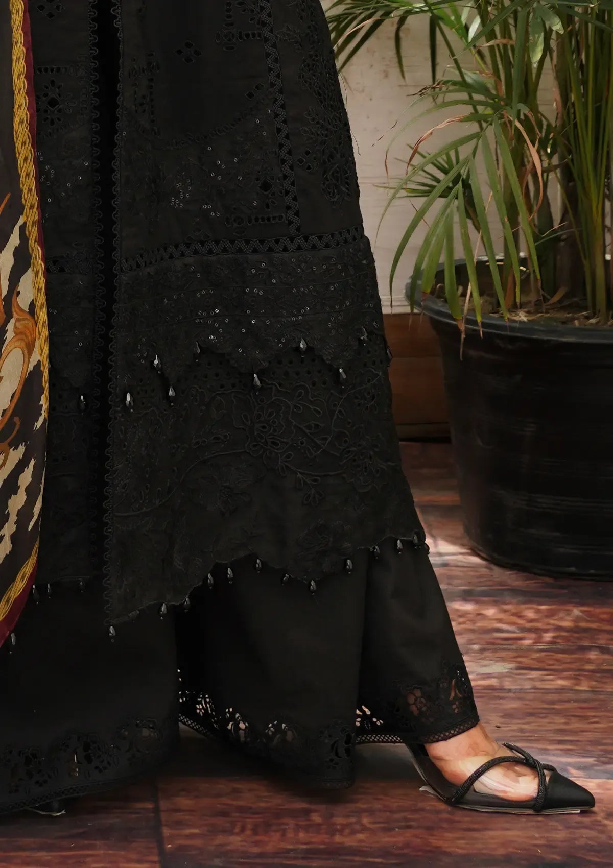 A close-up of a person wearing a black embroidered kurta and matching wide-leg trousers with lace trim, paired with black pointed flat sandals, standing on a wooden floor next to a potted plant.