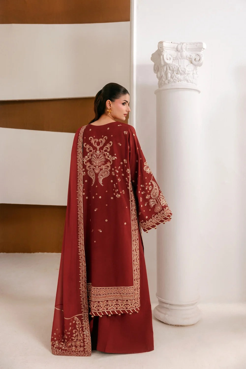 A woman wearing a deep maroon traditional outfit with intricate gold embroidery, standing next to a white decorative column in an indoor setting.