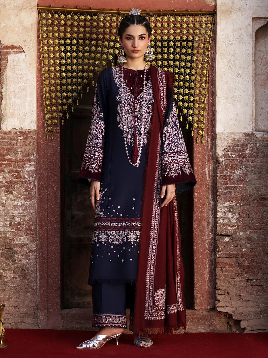 A woman models a traditional three-piece salwar kameez suit in navy blue and deep maroon, featuring intricate silver and white embroidery, set against a rustic stone wall and golden hanging curtain.