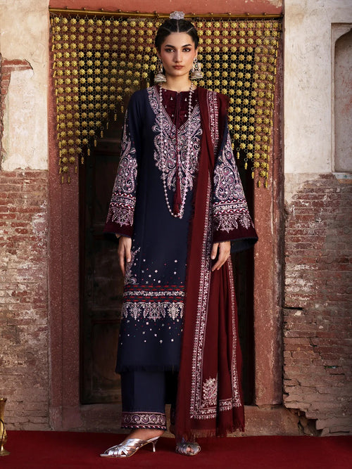A woman models a traditional three-piece salwar kameez suit in navy blue and deep maroon, featuring intricate silver and white embroidery, set against a rustic stone wall and golden hanging curtain.