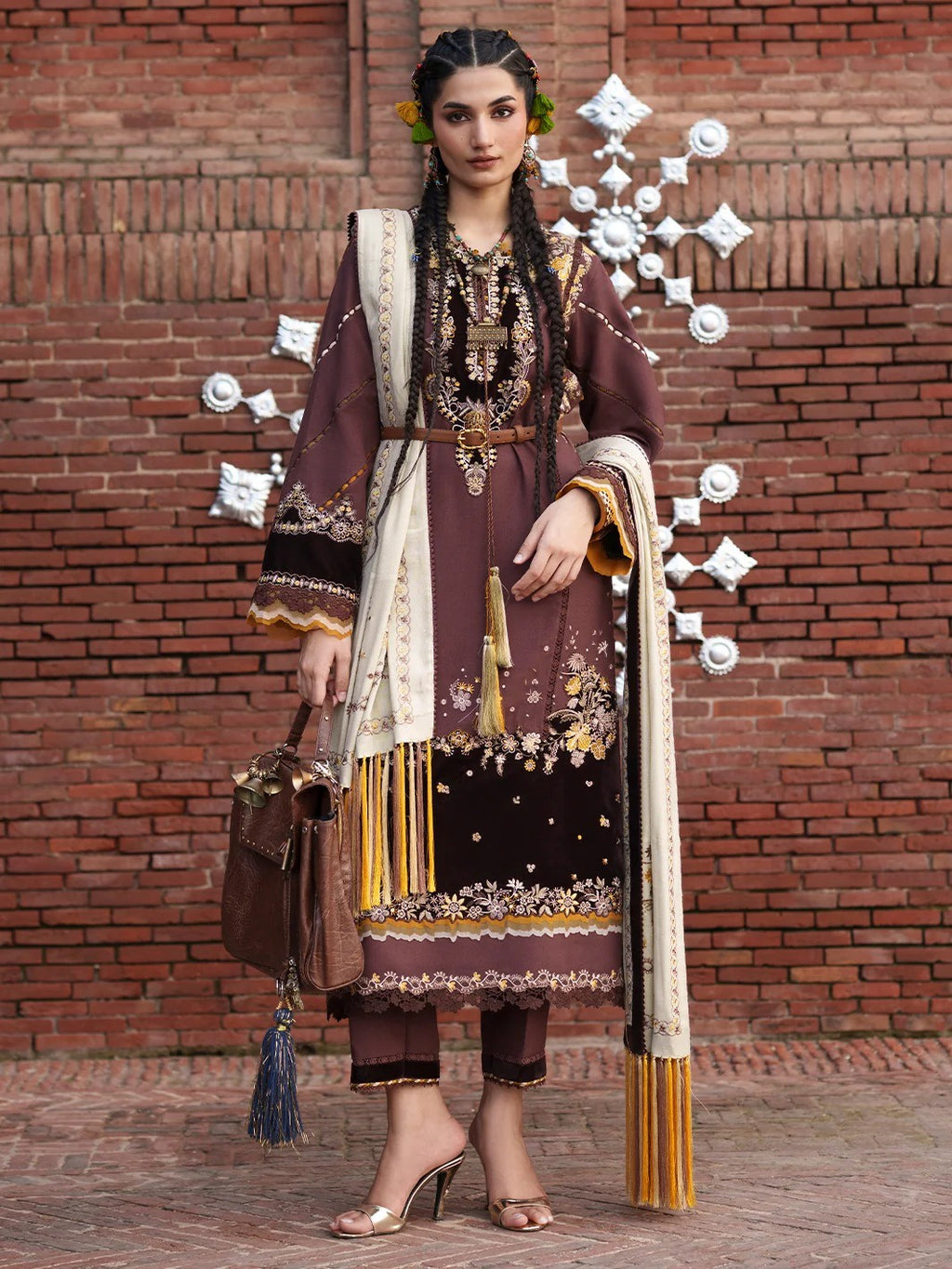 A woman models a traditional South Asian salwar kameez suit in a brown and gold colorway, featuring intricate embroidery, a cream dupatta, and a brown handbag, set against a red brick wall with decorative white metal ornaments.