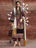 A woman models a traditional South Asian salwar kameez suit in a brown and gold colorway, featuring intricate embroidery, a cream dupatta, and a brown handbag, set against a red brick wall with decorative white metal ornaments.