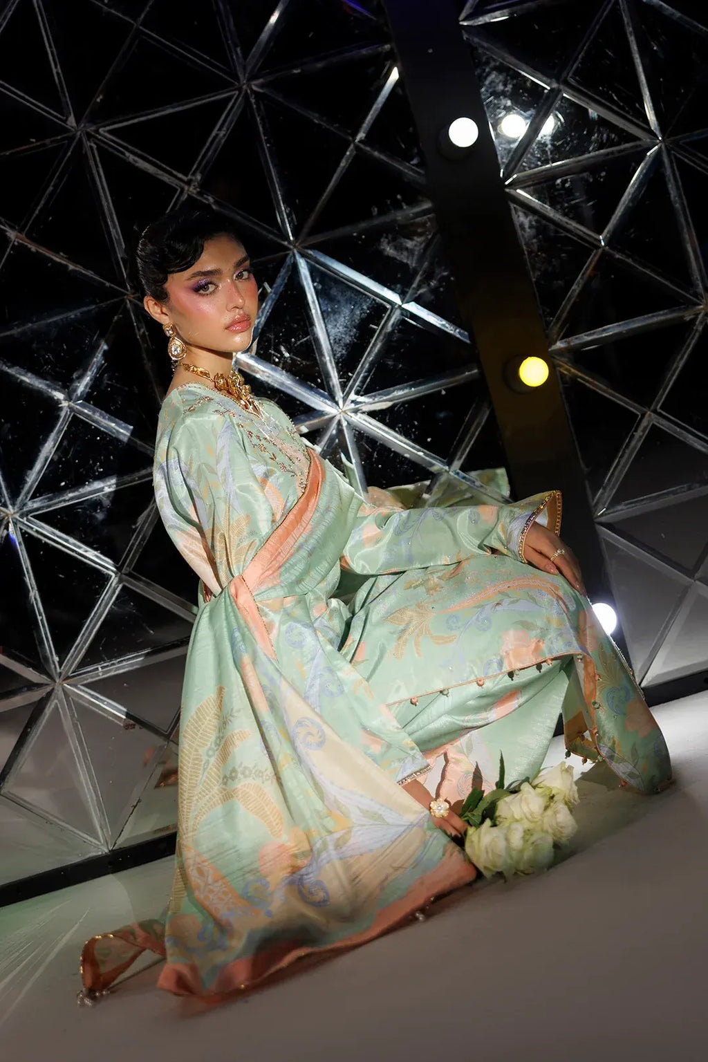 A woman models a traditional long-sleeved dress with a light mint green base and peach-pink and gold patterned details, posed in front of a modern geometric glass structure.