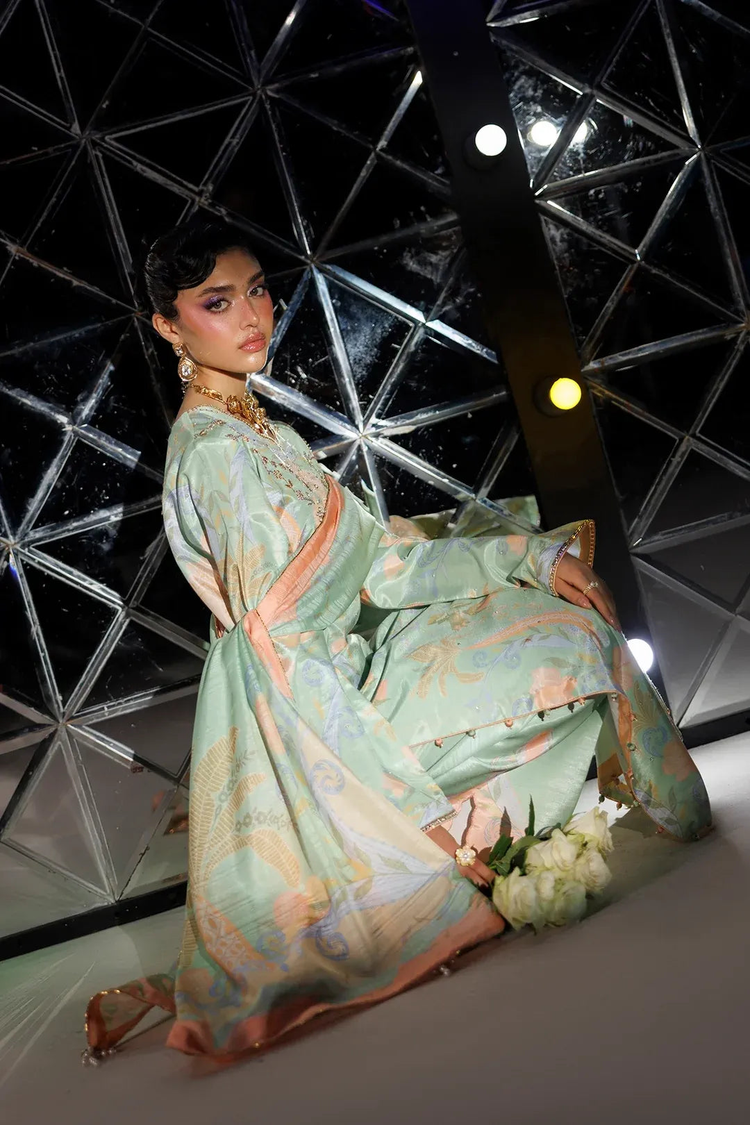 A woman models a traditional long-sleeved dress with a light mint green base and peach-pink and gold patterned details, posed in front of a modern geometric glass structure.