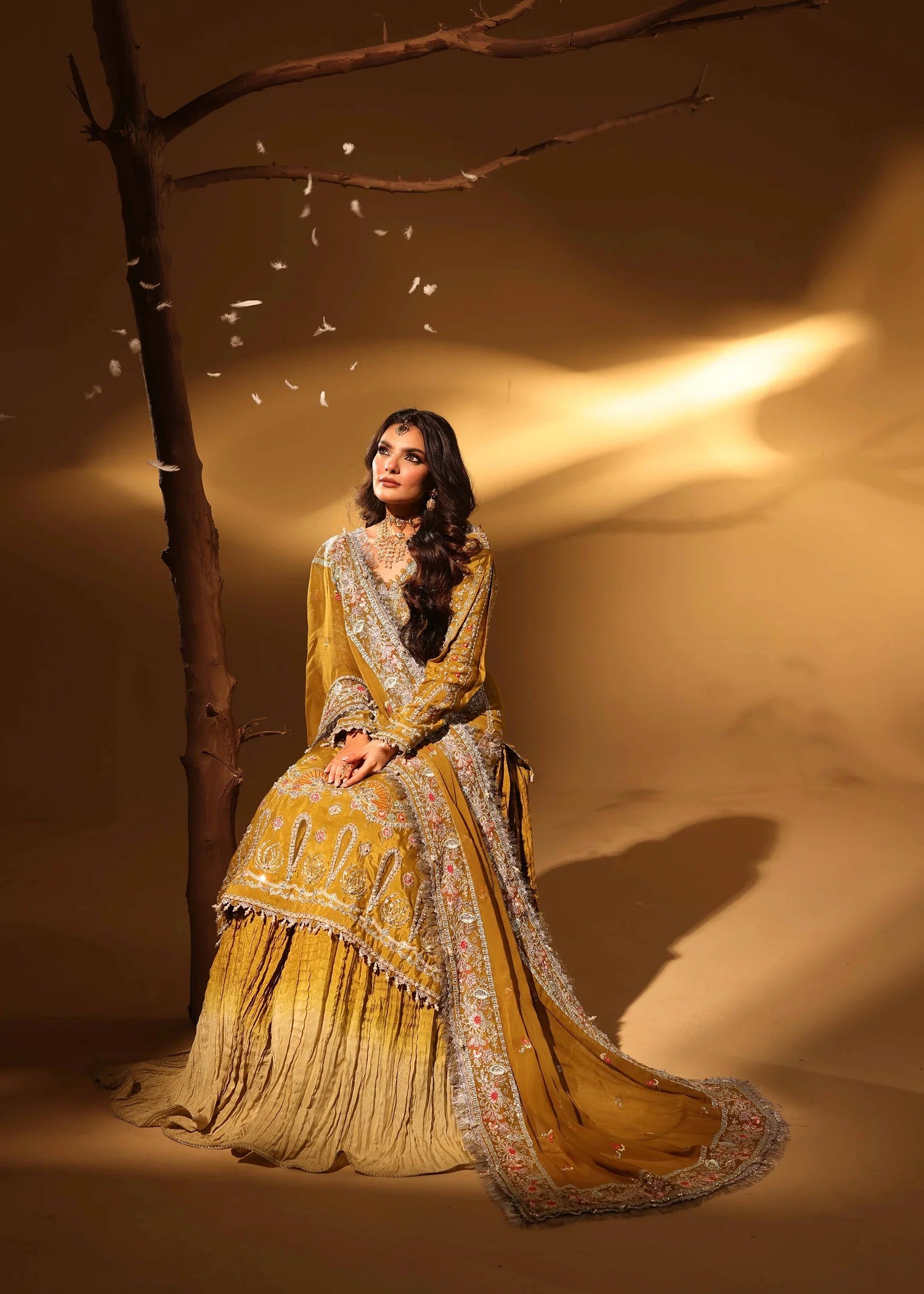 Woman in a traditional yellow and brown outfit standing against a warm-toned background with a tree branch.