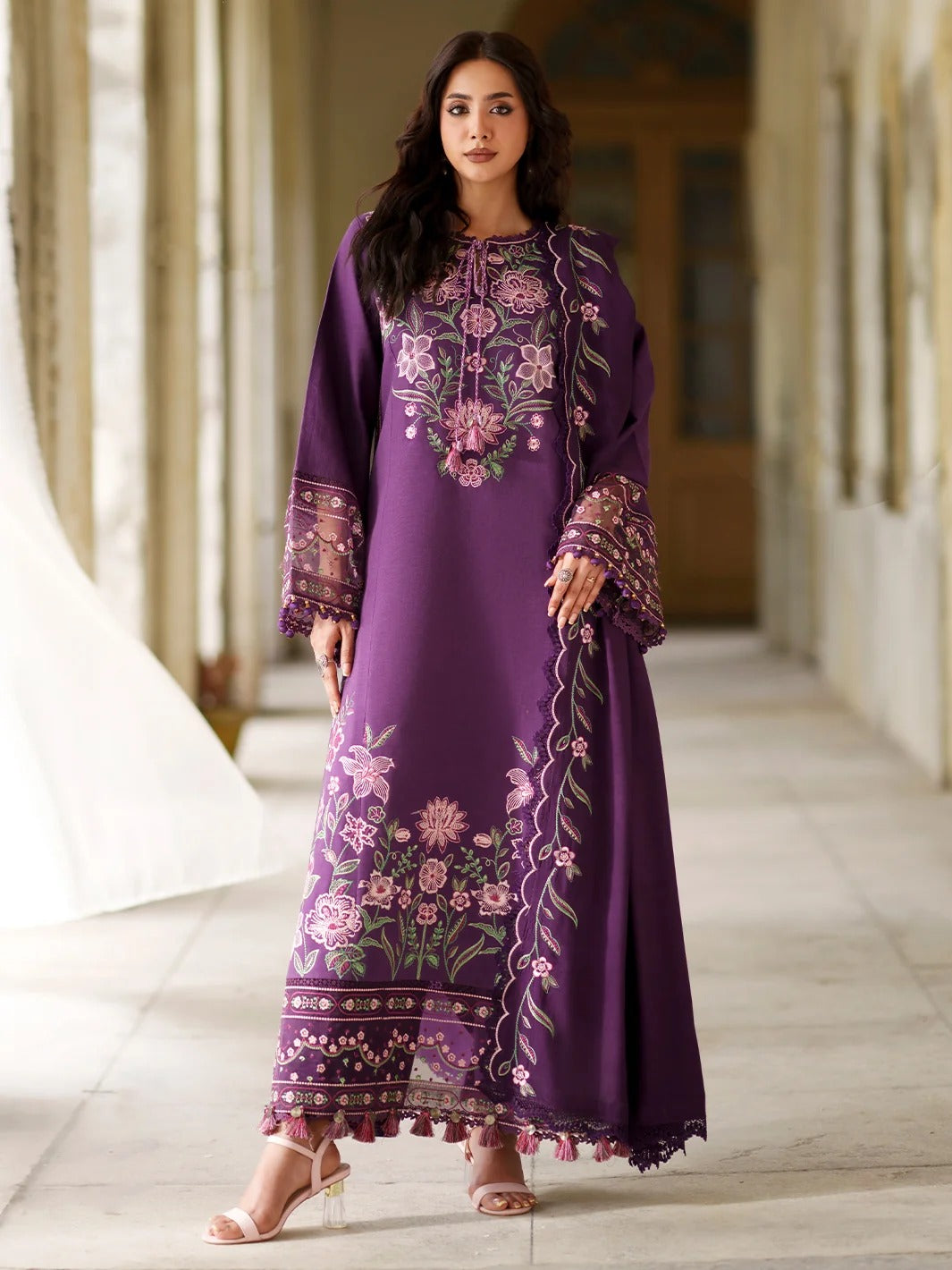A woman models a long purple embroidered kurta with floral patterns and tassel trim, standing in a corridor with columns.