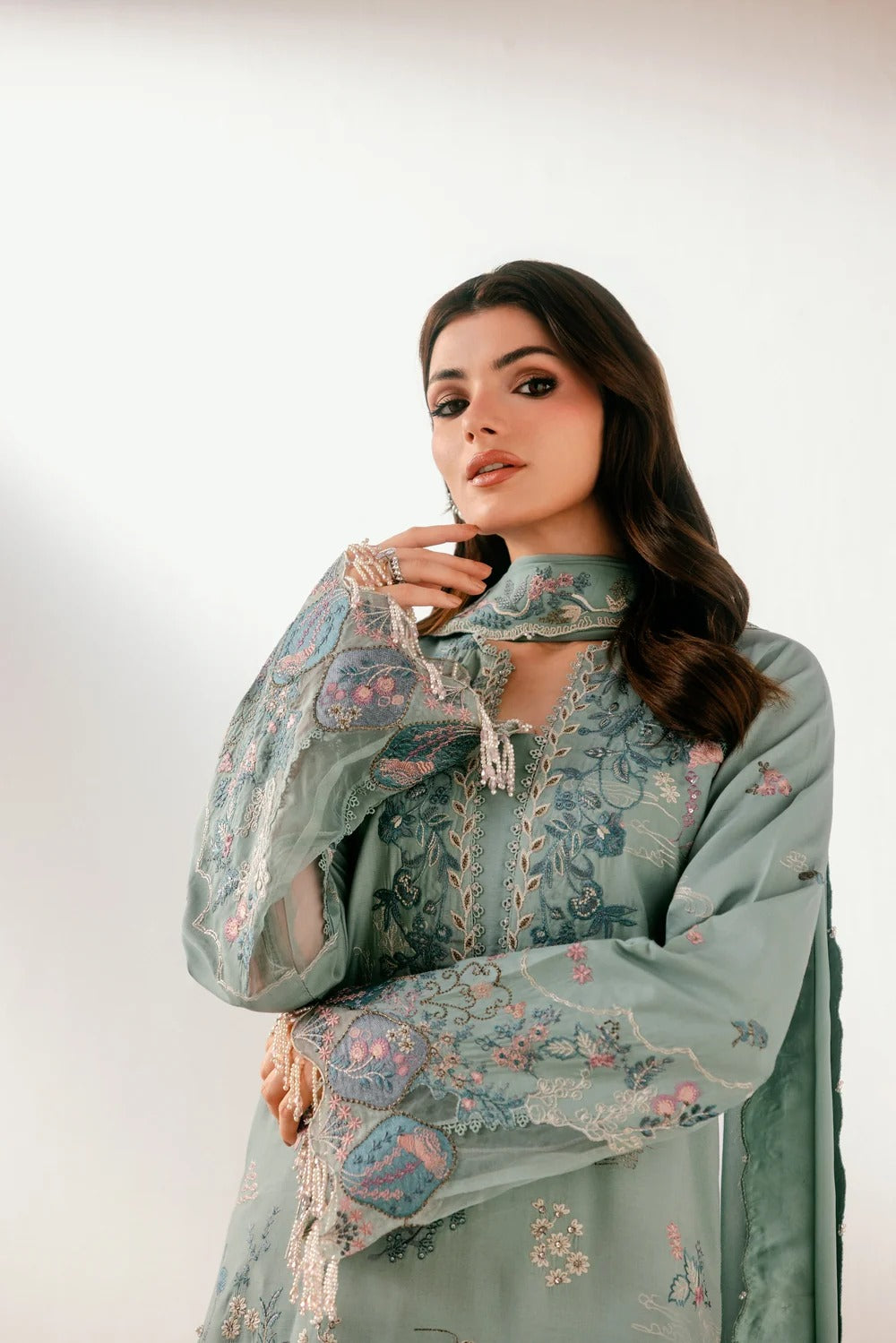 A woman modeling a traditional embroidered outfit in a sage green color with intricate floral patterns and lace details, set against a plain light background.