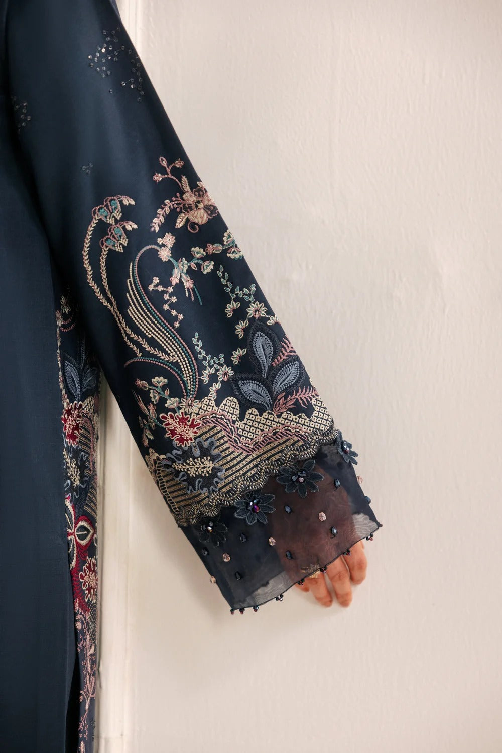 A close-up of a dark navy blue embroidered garment sleeve with intricate floral and bird motifs in beige, gold, and red, worn by a model whose hand is visible at the cuff.