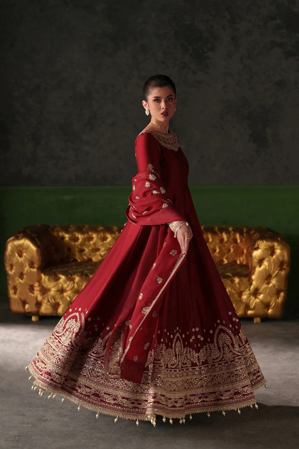 A woman models a traditional red Indian anarkali gown with intricate gold embroidery, standing in front of a gold tufted sofa against a dark textured wall.