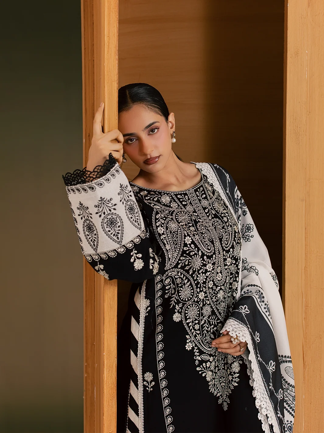 A woman models a black and white embroidered traditional outfit with a matching printed shawl, leaning against a wooden door frame.