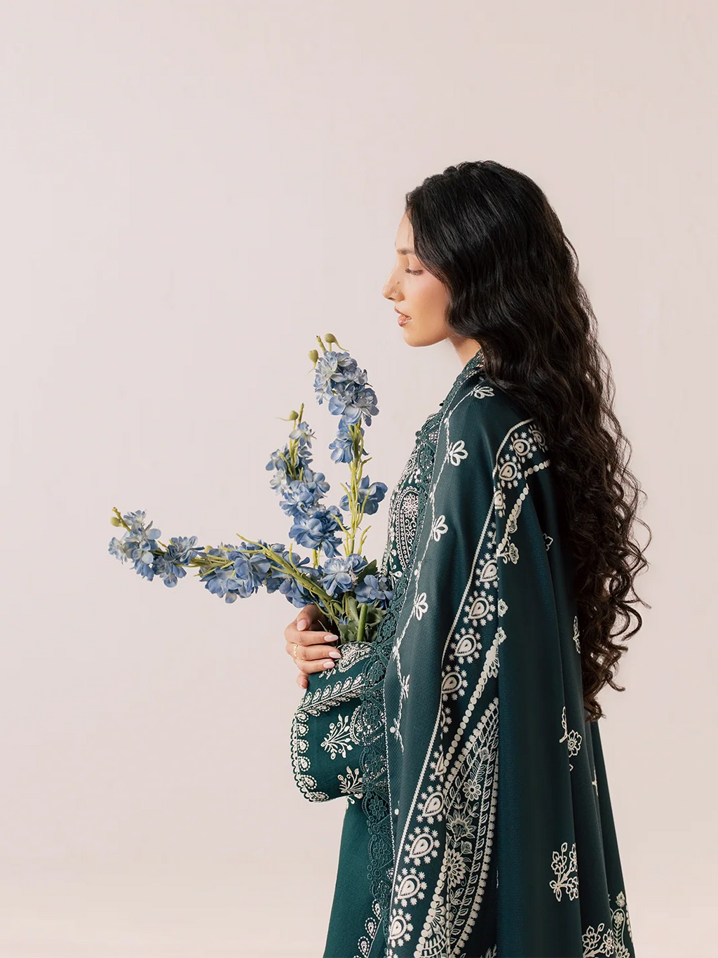 A woman in profile wearing a dark teal green embroidered shawl over a matching outfit, holding a bouquet of blue flowers against a plain light beige background.
