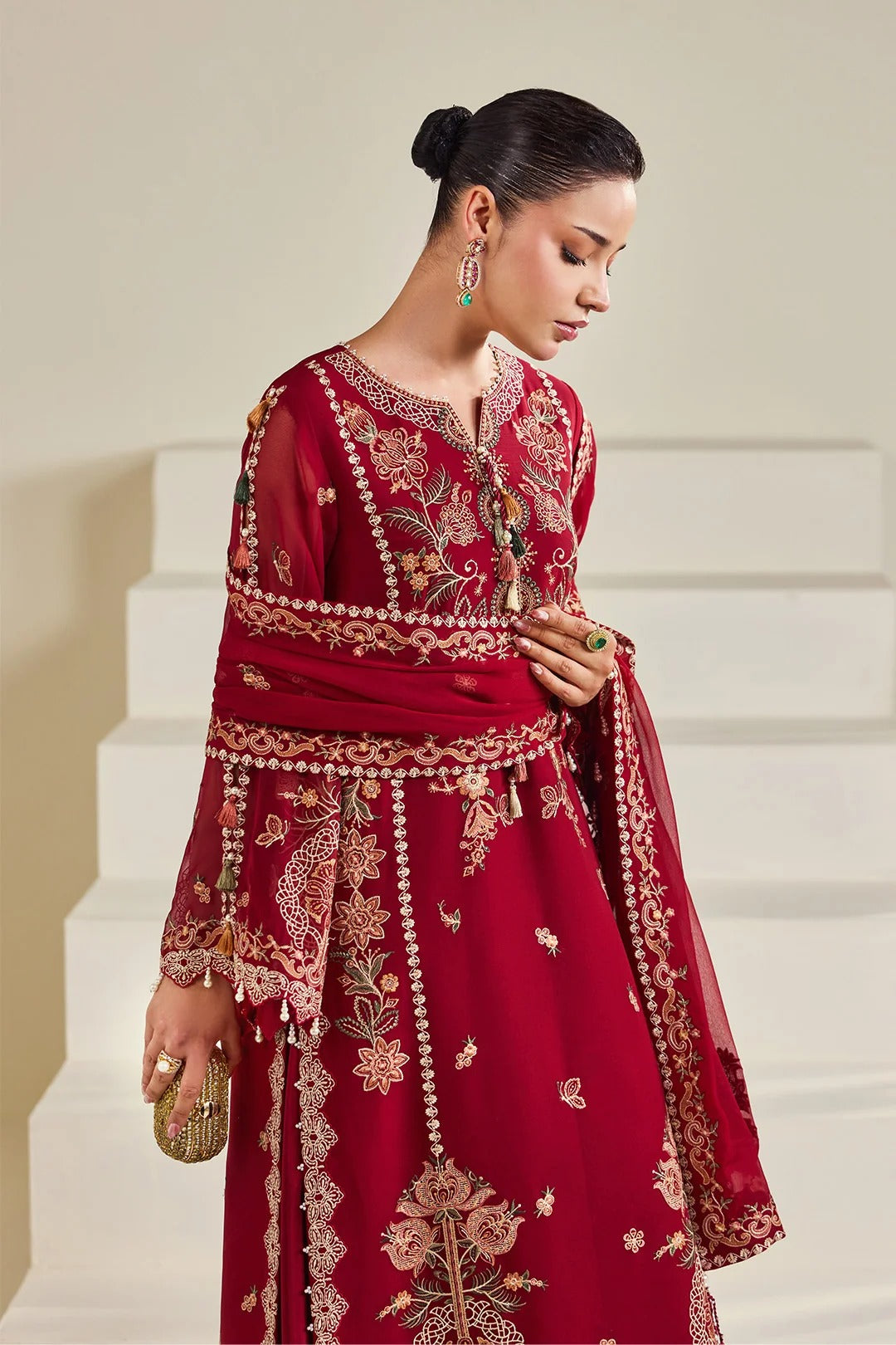 A woman models a deep red embroidered traditional outfit with a matching sheer dupatta, featuring intricate gold and beige floral embroidery, set against a minimalist beige staircase background.