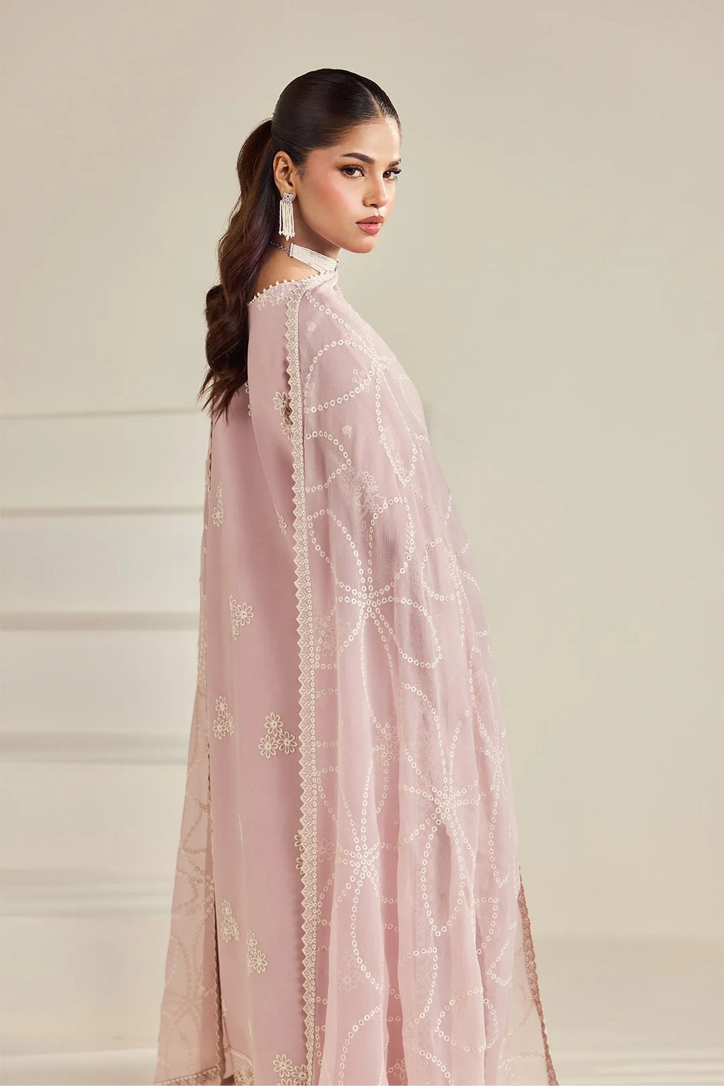 A woman wearing a light pink traditional outfit with a matching sheer embroidered dupatta, photographed against a soft beige studio background.